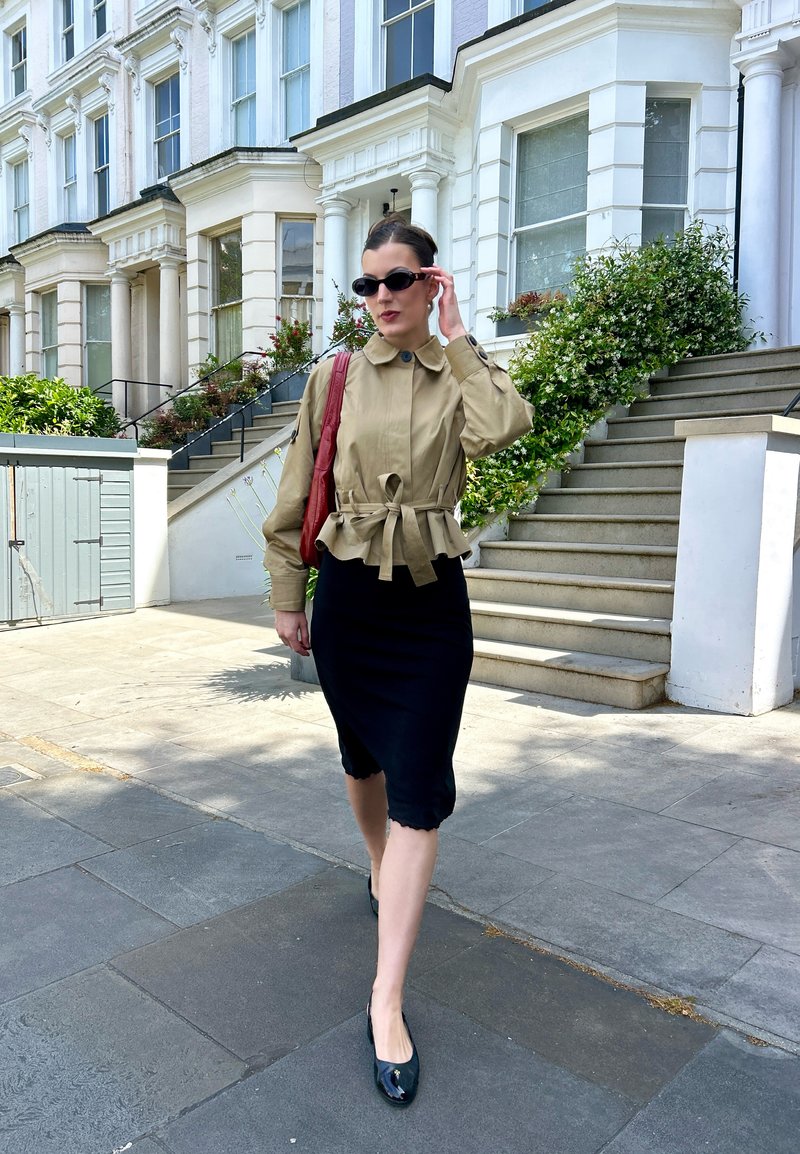 Femme portant des lunettes de soleil, une veste beige, une jupe noire et des chaussures noires marchant sur un trottoir à côté de maisons de ville blanches avec des escaliers et de la verdure.