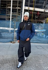 Νέος άνδρας που φοράει λευκό σκουφάκι, γυαλιά, μπλε-μαύρο μπουφάν Reebok, φαρδιά σκούρα τζιν και ασπρόμαυρα αθλητικά παπούτσια, στέκεται δίπλα σε εργοτάξιο.