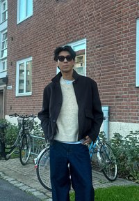 Crna vunena jakna s visokim ovratnikom, kremasti pleteni džemper i tamnoplave traperice širokih nogavica. Stojeći pored bicikala s pozadinom od ciglene zid.