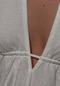 Bijela lanena odjeća s dubokim V-izrezom, nabrane teksture u struku i tankim vezicama za prilagodbu. Glatka tkanina, minimalni detalji.