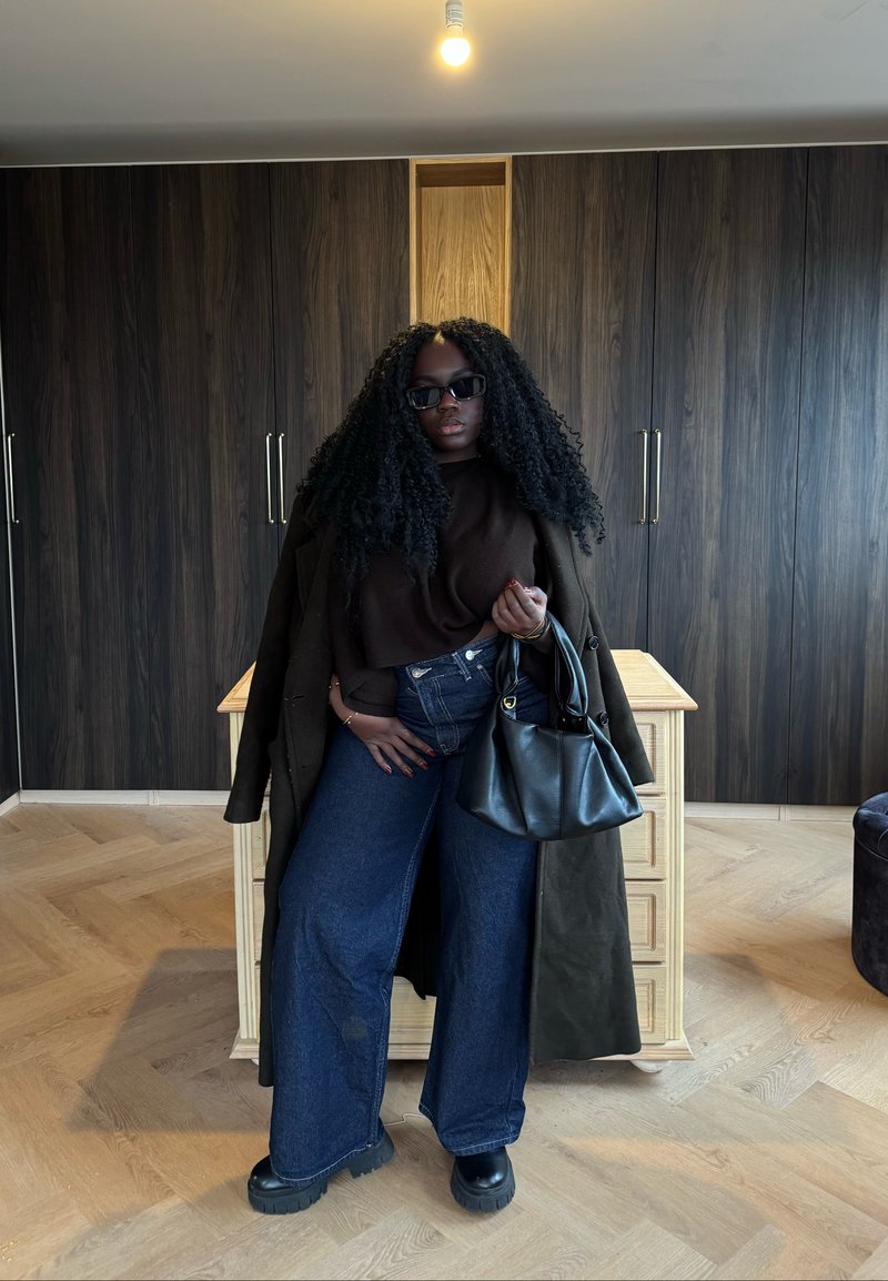 Personne aux cheveux longs et bouclés portant des lunettes de soleil, un manteau foncé, un haut marron, un jean à jambes larges, des bottes noires, tenant un sac à main noir dans une pièce moderne.