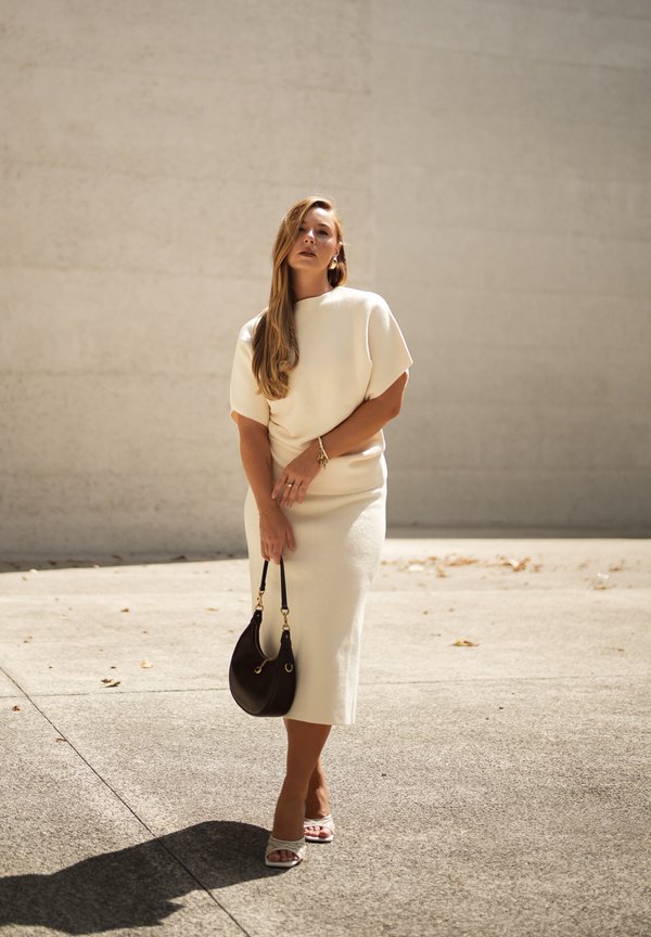 Femme en robe midi crème et sandales blanches tenant un sac à main noir, debout à l'extérieur devant un mur beige uni.