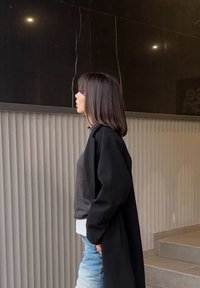 Mlada ženska s ramenoma dolgimi lasmi, oblečena v črni plašč in jeans hlače, stoji v profilu ob rebrasti steni in temni odsevni plošči.