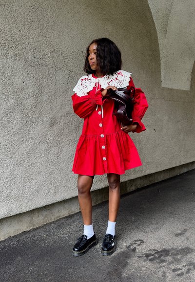 Vestido de algodón rojo con un gran cuello de encaje blanco, botones en la parte delantera y cintura fruncida. Zapatos negros y calcetines blancos completan el atuendo.
