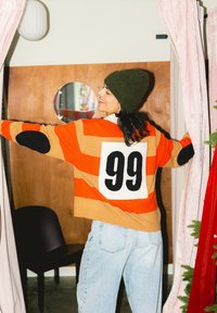 Femme portant un bonnet vert et un pull rayé orange avec le numéro 99 dans le dos, écartant des rideaux roses dans une pièce avec des boiseries.