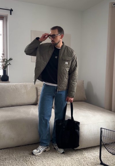 Hombre con gafas, chaqueta acolchada verde, camiseta negra, vaqueros azules, zapatillas blancas, sosteniendo un bolso tote negro en una sala de estar con un sofá beige.