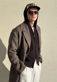 Homme portant des lunettes de soleil, casquette beige et noire, sweat à capuche zippé noir sous un manteau à carreaux, et pantalon de survêtement blanc, debout contre un mur beige.