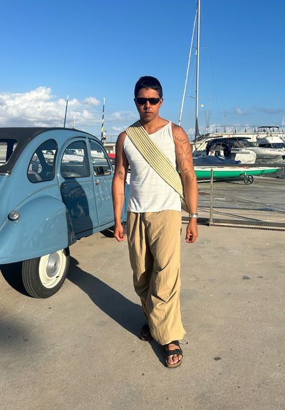 Top sin mangas de color gris claro, pantalones sueltos beige y sandalias negras. De pie junto a un coche vintage azul en un entorno de marina con barcos al fondo.