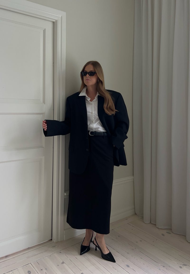 Femme portant un blazer noir surdimensionné, une chemise blanche, une jupe midi noire, des talons noirs et des lunettes de soleil, debout près d'une porte blanche et de rideaux à l'intérieur.