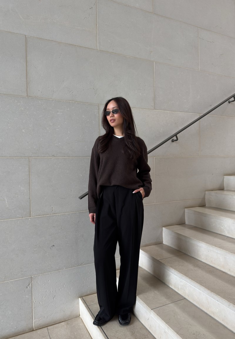 Femme portant un pull sombre, un pantalon noir large, des lunettes de soleil, debout à côté d’un escalier en pierre claire et d’un mur avec une rambarde en métal.