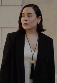 Blazer nero sopra una maglietta bianca, con una lunga collana che presenta una pendente rotonda di colore crema e un ponpon nero. Texture lisce e design semplice.