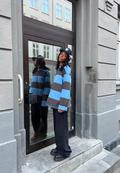 Camisa de rayas oversize en azul y marrón, pantalones oscuros, zapatos negros gruesos, de pie en una puerta con una superficie de vidrio reflectante.