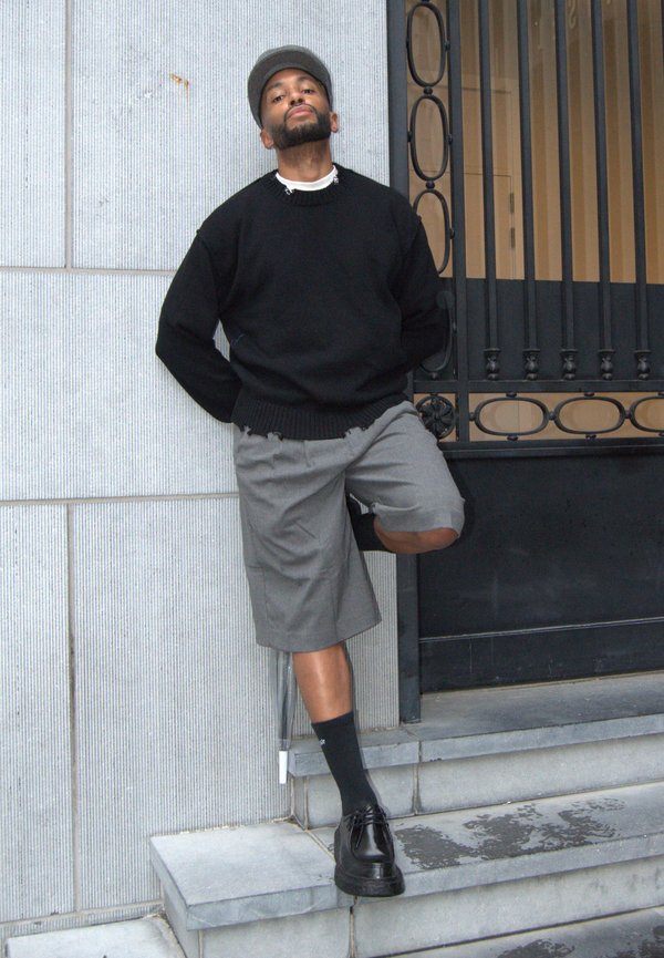 Schwarzer Pullover, graue Shorts, schwarze Socken und schwarze Schuhe. Person steht auf einer Stufe neben einem Tor mit vertikalen Streben. Beton Hintergrund.