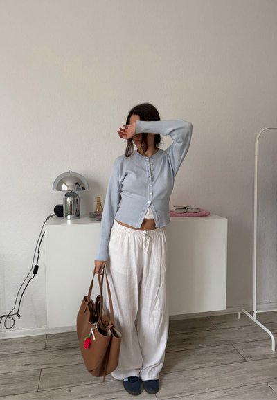 Mujer con cárdigan azul claro abotonado y pantalones blancos cubriéndose el rostro con el brazo, sosteniendo un bolso marrón, de pie en una habitación minimalista con lámpara y gafas.