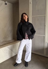 Femeie în picioare în interior, purtând o jachetă neagră supradimensionată, cămașă cu dungi, pantaloni largi albi, cizme negre, cu mâinile în buzunare și expresie neutră.