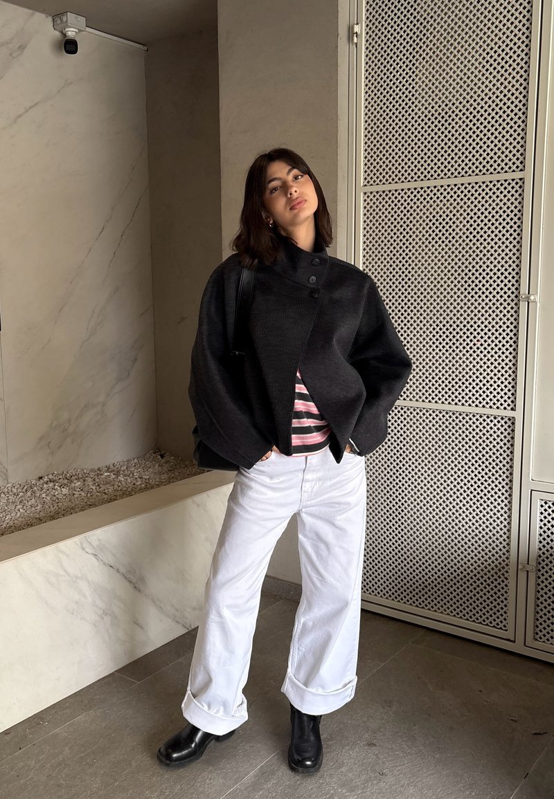 Femme debout à l'intérieur portant une veste noire surdimensionnée, une chemise rayée, un pantalon blanc à jambes larges, des bottes noires, les mains dans les poches, expression neutre.