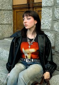 Μαύρο δερμάτινο μπουφάν, γραφικό κόκκινο και πορτοκαλί μπλουζάκι με σχέδιο φλόγας, ασημένια αλυσίδα ή necklace, φθαρμένα ανοιχτό μπλε τζιν, ζώνη με λοξά τρουκ.