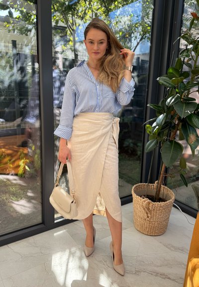 Camisa de botones a rayas azul claro y blanca, falda envolvente color crema, tacones altos beige y un pequeño bolso claro. Entorno interior con plantas.