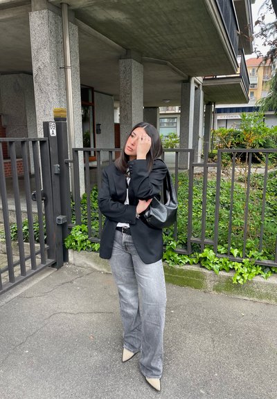 Americana negra oversize, camiseta gráfica blanca, jeans de pierna ancha gris y tacones beige de punta, con un bolso negro. Entorno de concreto.