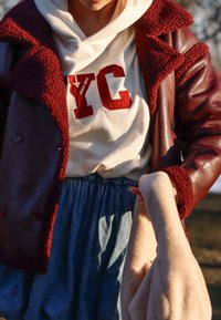 Bordeaux usnjen plašč z krznom obloženim notranjostjo, oblečen čez belo kapuco z rdečim napisom, v kombinaciji z svetlo modrim jeans krilom. Drži mehak, bež predmet.