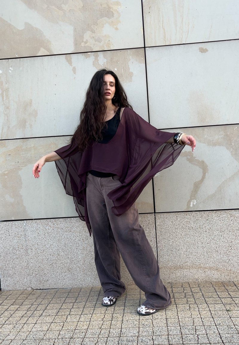 Donna con lunghi capelli scuri che indossa una blusa viola trasparente, pantaloni grigi larghi e scarpe a fantasia, in piedi contro un muro piastrellato con le braccia distese.