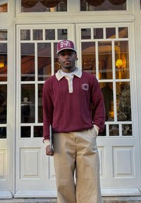 Homme portant un pull bordeaux avec un col blanc, un pantalon beige et une casquette, tenant un dossier devant des portes blanches à panneaux vitrés.