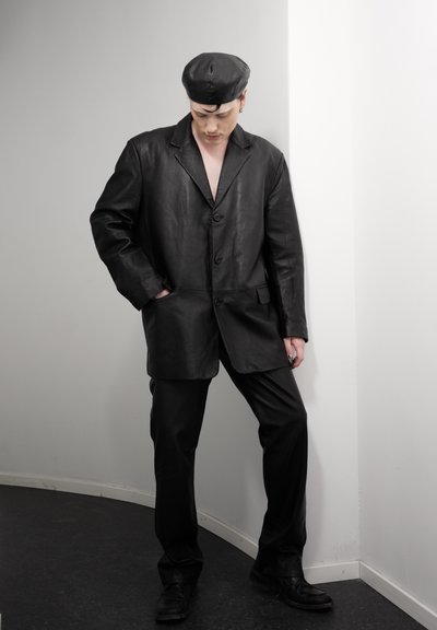 Persona con chaqueta de cuero negra, pantalones a juego y boina, de pie contra una esquina con una mano en el bolsillo, mirando hacia abajo.