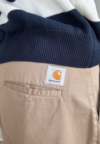 Pantalones Carhartt color beige con bolsillo trasero y parche de logo, combinados con un suéter de punto azul marino y blanco.