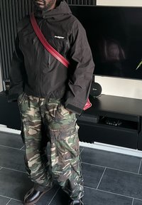 Veste imperméable noire avec capuche, pantalon cargo camouflage, et bottes marron. Un sac bandoulière rouge ajoute de la couleur à la tenue.
