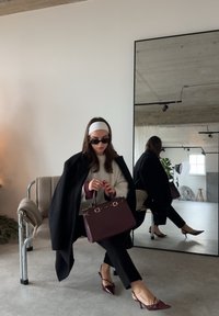 Femme en manteau noir et lunettes de soleil assise sur une chaise tenant un sac à main violet foncé, reflétée dans un grand miroir mural dans une pièce minimaliste.