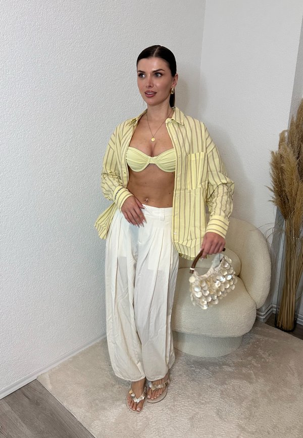 Vrouw draagt een geel gestreept shirt over een geel bandeau, witte wijde broek, houdt een witte schelptas vast, staand bij een beige stoel en gedroogde planten.