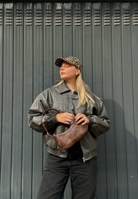 Braune Lederhandtasche mit geschwungenem Design, kombiniert mit einer grauen oversized Jacke aus Kunstleder und einer Leopardenmuster-Mütze, vor einem grauen Hintergrund.
