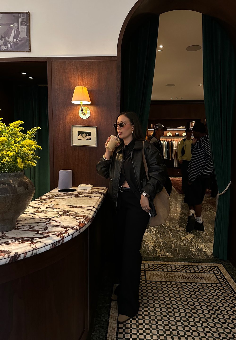 Must mustriga jope musta toppi peal, tumedad prillid, käes jook. Marmorist lett koos lillekimpuga. Rohelised kardinad taustal.