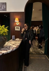 Must mustriga jope musta toppi peal, tumedad prillid, käes jook. Marmorist lett koos lillekimpuga. Rohelised kardinad taustal.