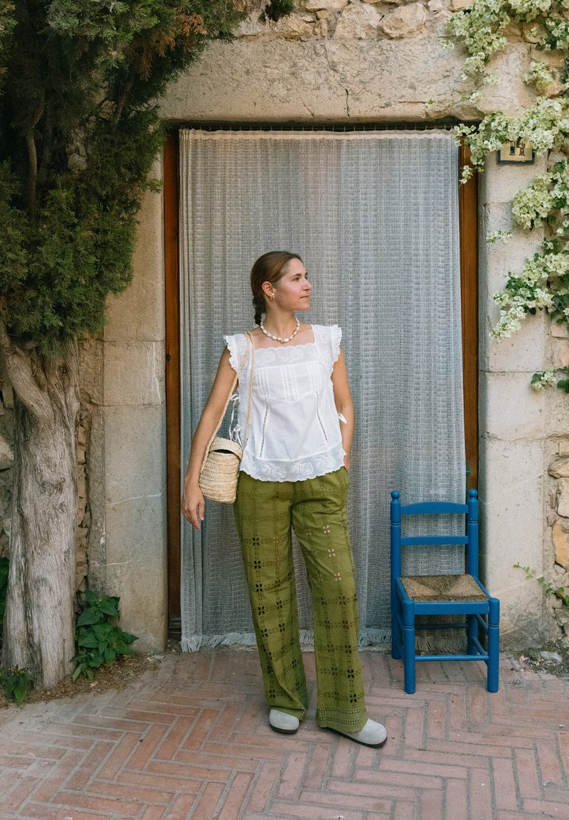 Γυναίκα με λευκό πουκάμισο και πράσινο παντελόνι στέκεται κοντά σε πετρόχτιστο τοίχο με πόρτα καλυμμένη με κουρτίνα και μπλε ξύλινη καρέκλα σε εξωτερικό χώρο.