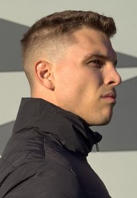 Μαύρο μπουφάν με ψηλό γιακά, με υφή ύφασμα. Κοντινό πλάνο προφίλ με κοντά, υφασματοποιημένα μαλλιά και ουδέτερη έκφραση σε γκρι φόντο.