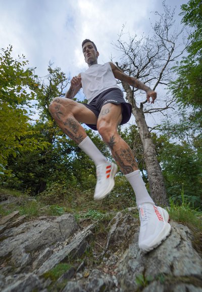 Zapatos deportivos blancos con acentos rojos, usados con calcetines blancos, en un terreno rocoso. La persona lleva una camiseta sin mangas y pantalones cortos, rodeada de vegetación.