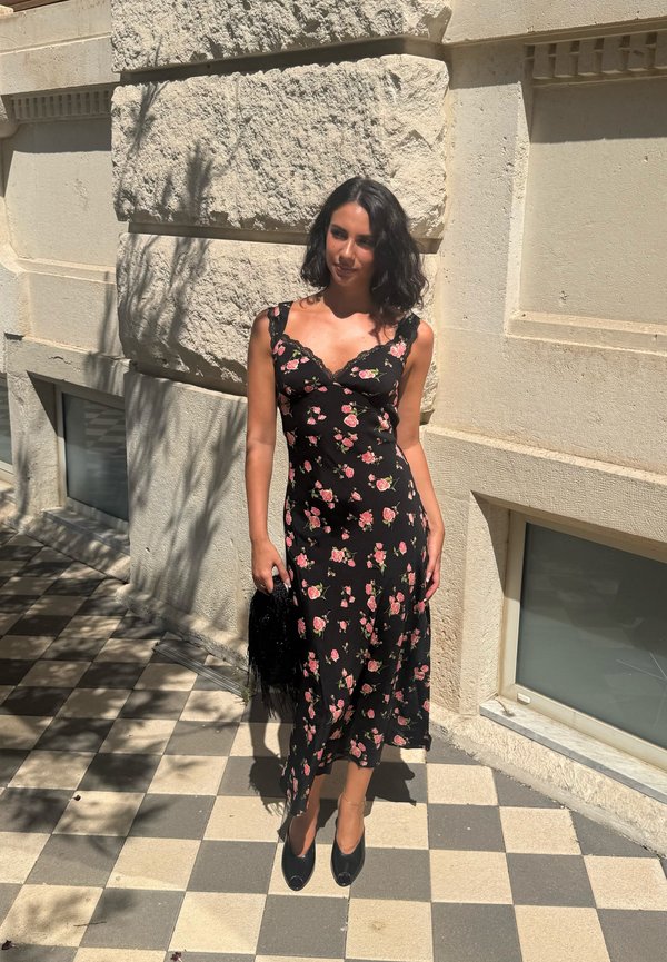 Femme aux cheveux noirs portant une robe noire fleurie et des chaussures noires se tient sur un sol à carreaux devant un mur de pierre.
