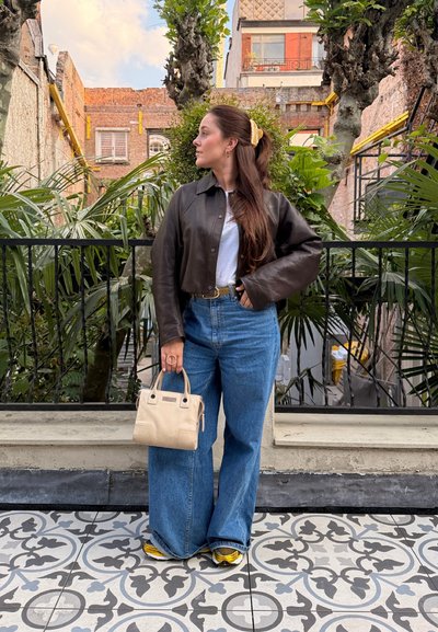 Mujer con el cabello largo y castaño que lleva una chaqueta de cuero negra, vaqueros anchos azules y zapatillas amarillas, está de pie sosteniendo un bolso beige en un área de jardín al aire libre.
