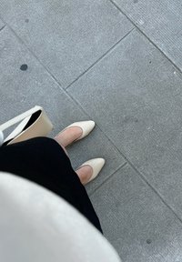 Des chaussures à talons hauts en cuir beige avec un bout pointu, assorties à une robe noire, et un sac à main de couleur claire sur un pavé gris texturé.