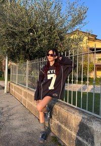 Black oversized sweatshirt featuring a white "NYU 7" print, teamed with grey trainers and knee-high socks, standing against a stone wall.