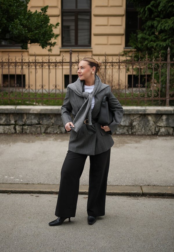 Vrouw met een grijze blazer en een gestrikte trui, zwarte broek met brede pijpen en laarzen met hakken, staand op een stoep naast een stenen muur en een hek.