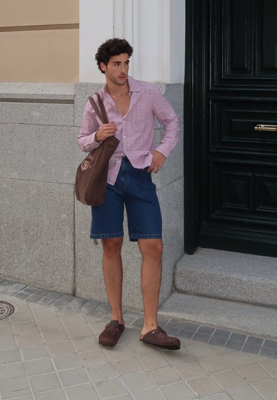 Camisa de botones a rayas rosas y blancas, pantalones cortos de mezclilla azules, zapatos marrones sin cordones y una bolsa de mano marrón con logo. De pie contra una pared.