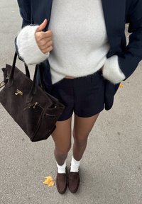 Marineblauwe pinstripe blazer over een witte fuzzy trui, donker suède handtas, marineblauwe shorts, witte sokken en bruine suède instappers op een verharde ondergrond.
