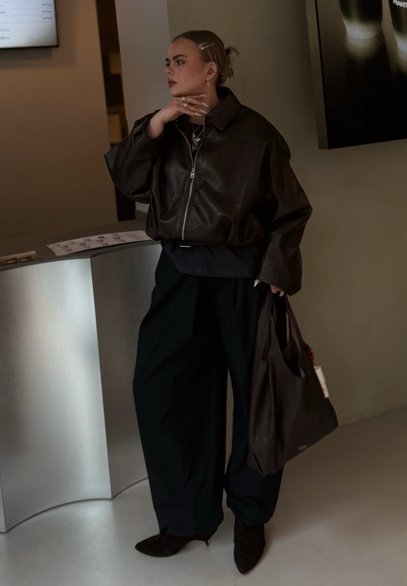 Persoană tânără cu părul dat pe spate poartă o geacă din piele neagră supradimensionată, pantaloni negri largi, ținând o geantă de mână în interior, lângă un tejghea metalic curbat.