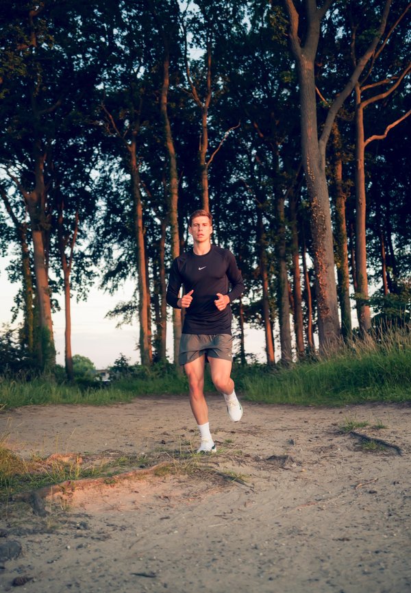 Corredor con una camiseta negra de manga larga y pantalones cortos grises, llevando zapatillas blancas, en un camino de arena con árboles al fondo durante el atardecer.