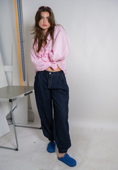 Mujer joven con cabello largo y castaño, vistiendo una blusa rosa claro, pantalones oscuros y sueltos, zapatos azules tipo slip-on y gafas de sol rosas en interiores.