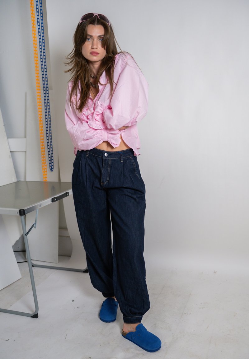 Giovane donna con capelli castani lunghi, che indossa una camicetta rosa chiaro, pantaloni scuri larghi, scarpe blu slip-on e occhiali da sole rosa all'interno.