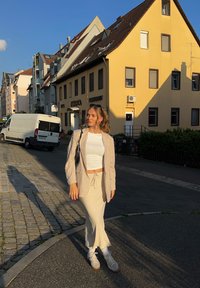 Beżowa lniana marynarka na białym topie crop i jasnej spódnicy, zestawiona z białymi skarpetkami i jasnymi trampkami. W tle kamienista ulica i budynki.