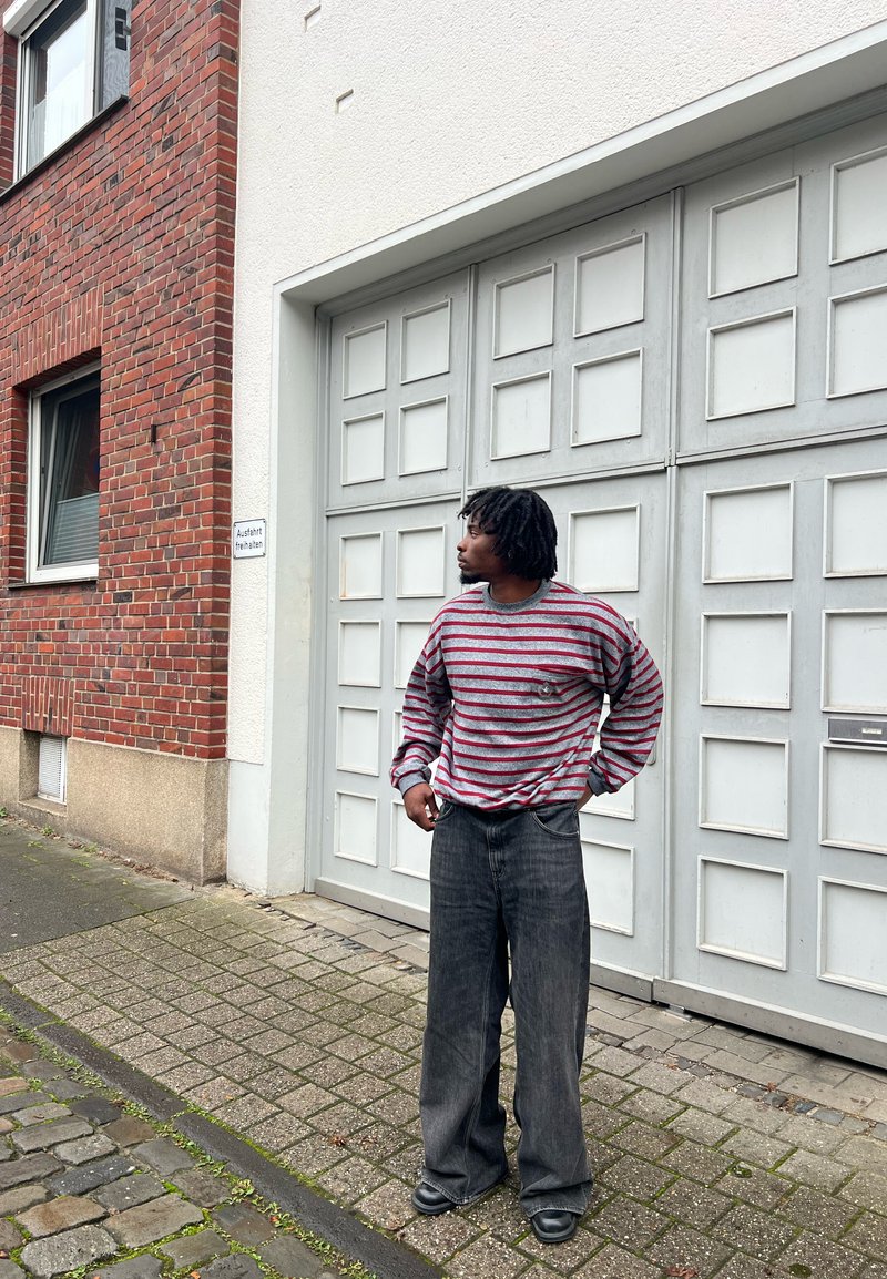 Gestreepte grijze en rode langslavige shirt met een ronde hals, gecombineerd met wijde zwarte denim broek. Zwarte schoenen, staande nabij een stenen muur.